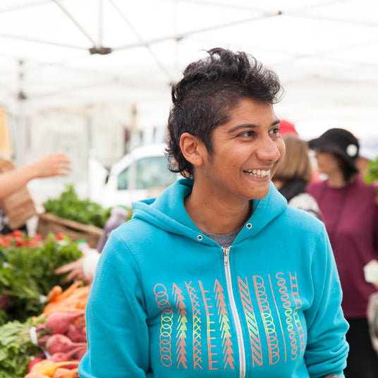 A young person wearing a bright blue ""Burlap & Barrel"" hoodie smiles at a farmers market with fresh produce visible in the background.