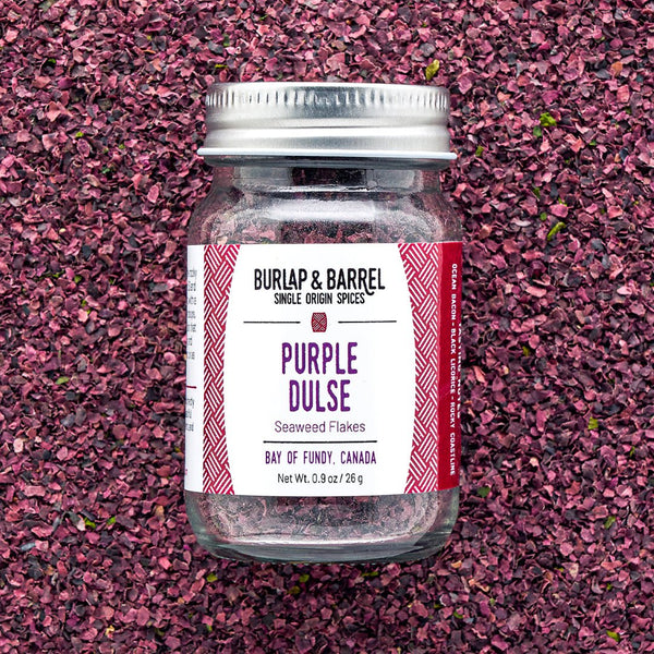 A jar of Purple Dulse Flakes by Burlap & Barrel, sustainably hand-harvested from the Bay of Fundy, Canada, displayed on a background of dulse flakes.