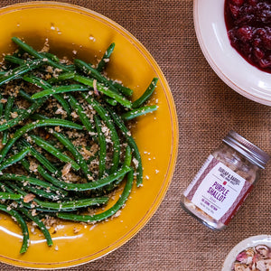 Yellow plate with green beans, jar of Burlap & Barrel purple shallot slices, and bowl of cranberries on a brown textured surface.