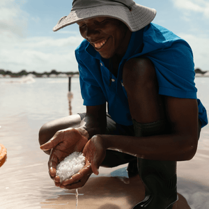 Partner farmer smiling in a blue shirt and tan hat, holding two handfuls of hand harvested pearl salt in Tanzania. 