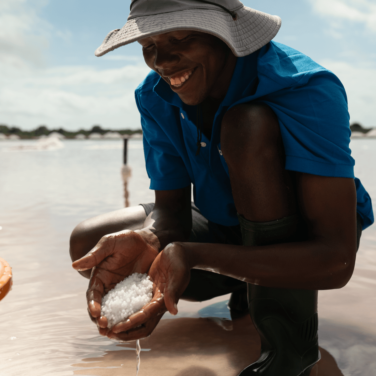 Partner farmer smiling in a blue shirt and tan hat, holding two handfuls of hand harvested pearl salt in Tanzania. 
