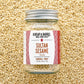 A glass jar of Sultan Sesame seeds by Burlap & Barrel, filled with sesame seeds and topped with a silver metal lid, set against a background of loose sesame seeds. The label features a white background with red patterned borders and brown text.