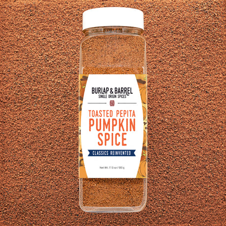 A clear plastic spice jar of Burlap & Barrel Toasted Pepita Pumpkin Spice with a white lid and white label, set against a textured brown background.
