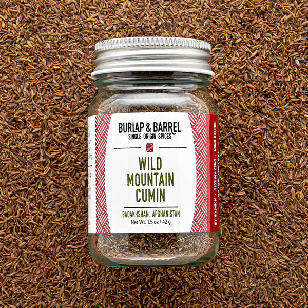 Glass jar of wild mountain cumin from Burlap & Barrel with a metal lid on a background of loose cumin seeds.