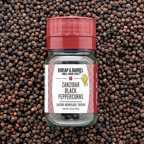 A glass jar of Zanzibar Black Peppercorns from Burlap & Barrel with a red lid, set against a background of loose black peppercorns.
