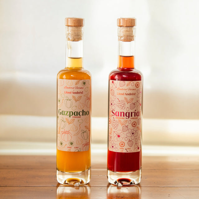 Two clear glass bottles of natural liquid nectaries with cork stoppers, standing on a wooden table in front of a softly blurred window. The left bottle is labeled Gazpacho with golden yellow liquid, and the right bottle is labeled Sangrie with deep red liquid.