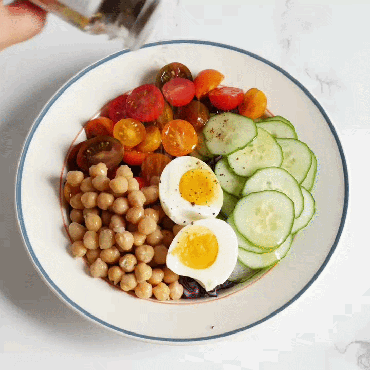 lemon sumac pepper getting sprinkled on chickpeas, hard boiled egg, cucumber and cherry tomatoes in a white bowl with a blue rim. 