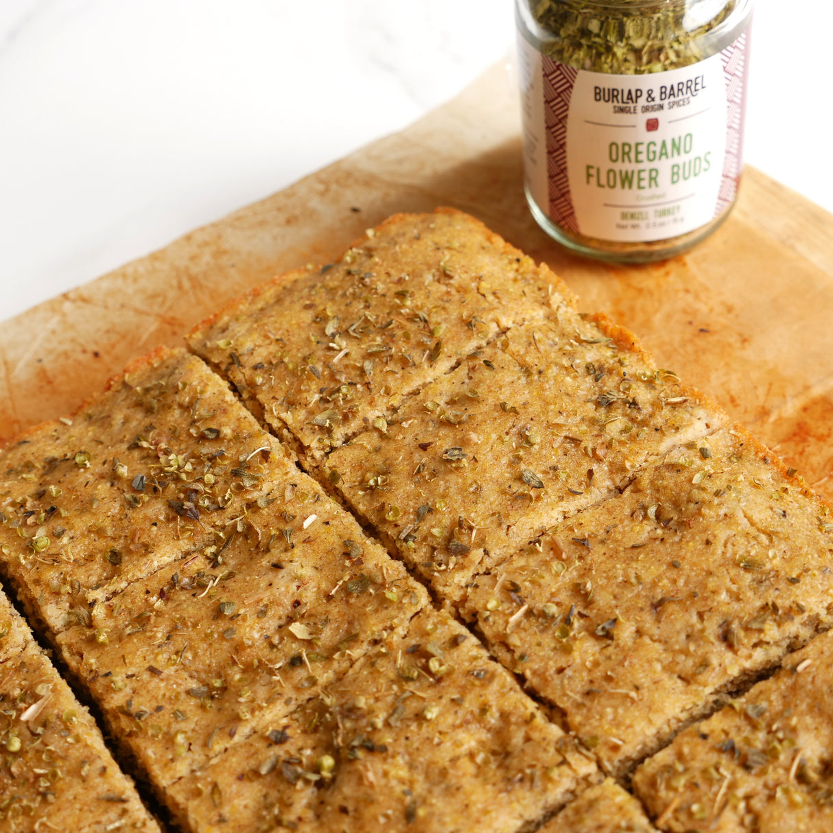Square slices of bread with oregano on a wooden board next to a jar of oregano.