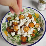 Cucumber salad with orange cherry tomatoes and feta crumble on a white plate with painted green leaves on the edge.