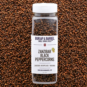 A plastic container of Zanzibar Black Peppercorns from Burlap & Barrel with a white lid, set against a background of loose black peppercorns.